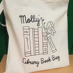 May include: White canvas tote bag with black text and a graphic of a stack of books and a butterfly. The text reads "Molly's Library Book Bag".