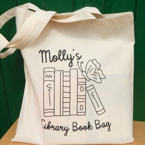 May include: A white canvas tote bag with black text and a graphic of books and a butterfly. The text reads "Molly's Library Book Bag".