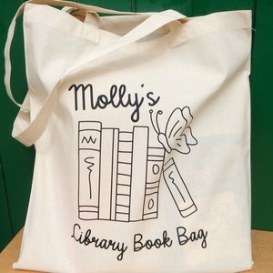 May include: A white canvas tote bag with black text and a graphic of a stack of books and a butterfly. The text reads "Molly's Library Book Bag".