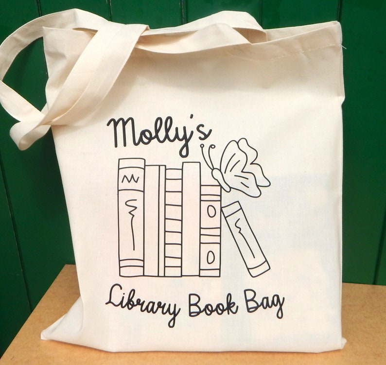 May include: A white canvas tote bag with black text and a graphic of a stack of books and a butterfly. The text reads "Molly's Library Book Bag".