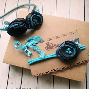 May include: Two hair accessories with blue and black fabric flowers. The headband has a turquoise elastic band and the barrette has a turquoise ribbon.