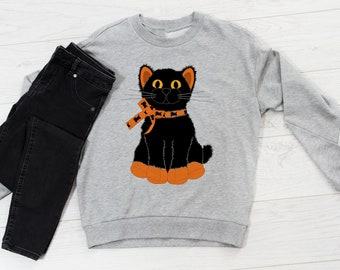 May include: A gray sweatshirt with a black cat wearing an orange and black scarf. The cat has yellow eyes and orange paws. The sweatshirt is laying on a white wooden surface with a pair of black jeans.
