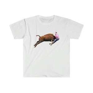 Może przedstawiać: Biały t-shirt z grafiką przedstawiającą mężczyznę jeżdżącego na brązowym bizonym. Mężczyzna ma na sobie różową koszulę i niebieskie dżinsy.
