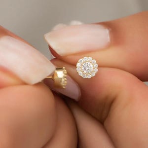 May include: A gold stud earring with a round, clear center stone surrounded by smaller clear stones.