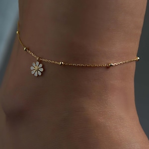 May include: A gold chain anklet with a small white daisy charm. The chain is made of small gold beads.