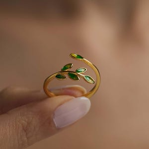 Könnte beinhalten: Ein goldener Ring mit einem zarten grünen Blattdesign. Die Blätter sind in einer astähnlichen Form angeordnet und erzeugen einen natürlichen und eleganten Look.