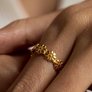 Puede incluir: Un anillo dorado con un diseño floral repetido. El anillo está hecho de un metal brillante y se usa en un dedo. Las pequeñas flores detalladas crean un diseño delicado. El diseño del anillo es femenino y elegante.