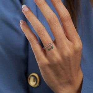 May include: A silver ring with a delicate branch design and two green leaves. The ring is worn on a finger with white nail polish.