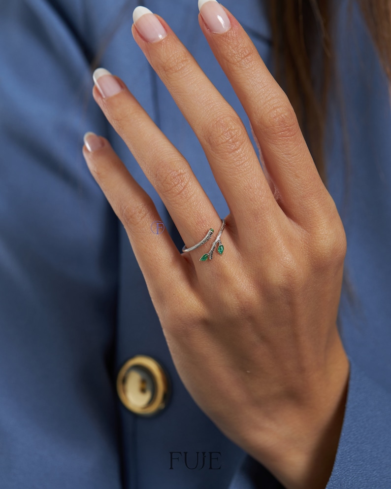 May include: A silver ring with a delicate branch design and two green leaves. The ring is worn on a finger with white nail polish.