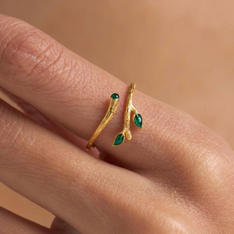 May include: A gold ring with a twig design and three green leaves. The ring is open and wraps around the finger.