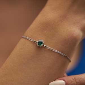 May include: A delicate silver chain bracelet featuring a small, round green gemstone encircled by a halo of smaller clear gemstones. The bracelet is worn on a person's wrist.