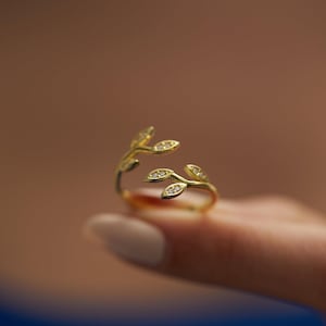 May include: A gold ring featuring a delicate design of two branches with leaves, each leaf set with a small clear stone.