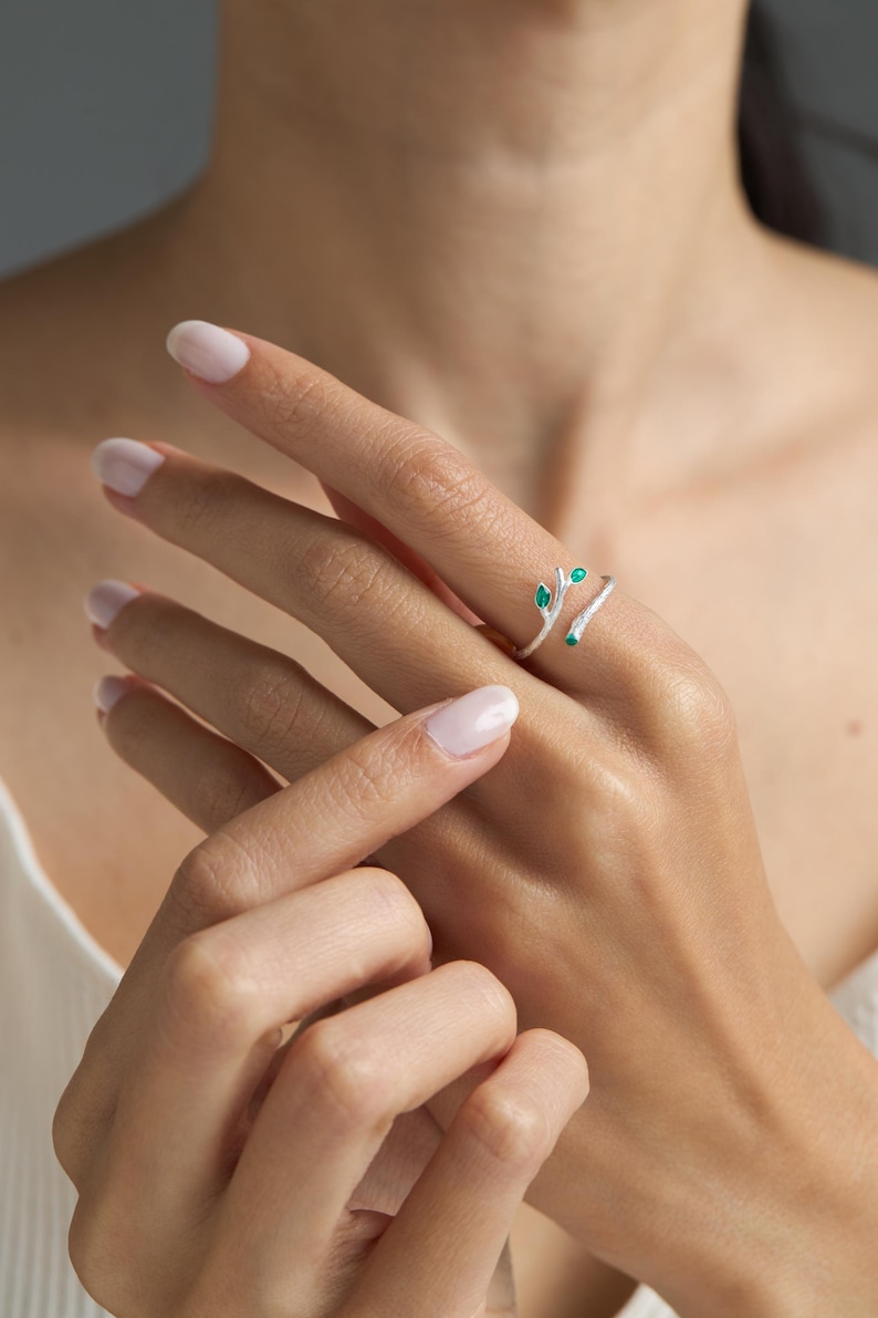 May include: A silver ring with a delicate branch design and green gemstone accents. The ring is open and wraps around the finger.