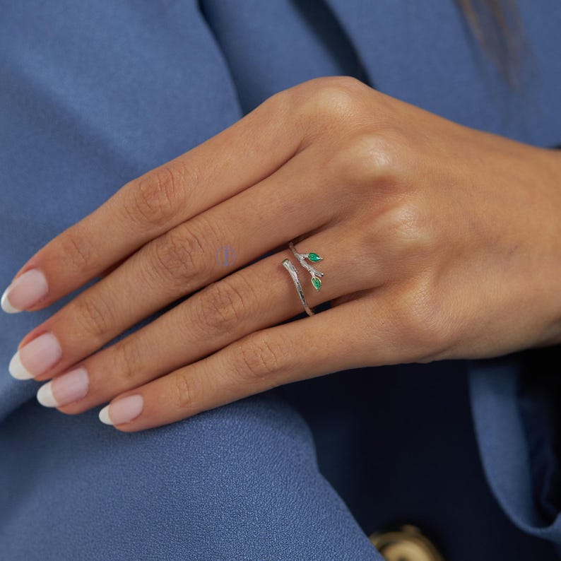 May include: A silver ring with a delicate branch design and two green gemstones. The ring is worn on a finger.