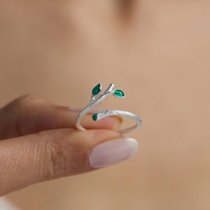 May include: A silver ring with a twig design and three green leaves. The ring is open and wraps around the finger.