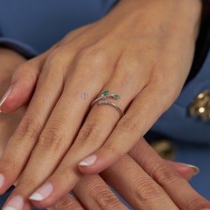 May include: A silver ring with a delicate branch design and two green gemstones. The ring is worn on a person's finger.