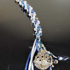 May include: A beaded keychain with a silver and blue color scheme. The keychain has a silver charm with a floral design and a tassel with blue and white ribbons.