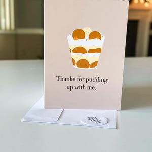 May include: A light brown card with a cartoon illustration of a banana pudding dessert. The text on the card reads "Thanks for pudding up with me."