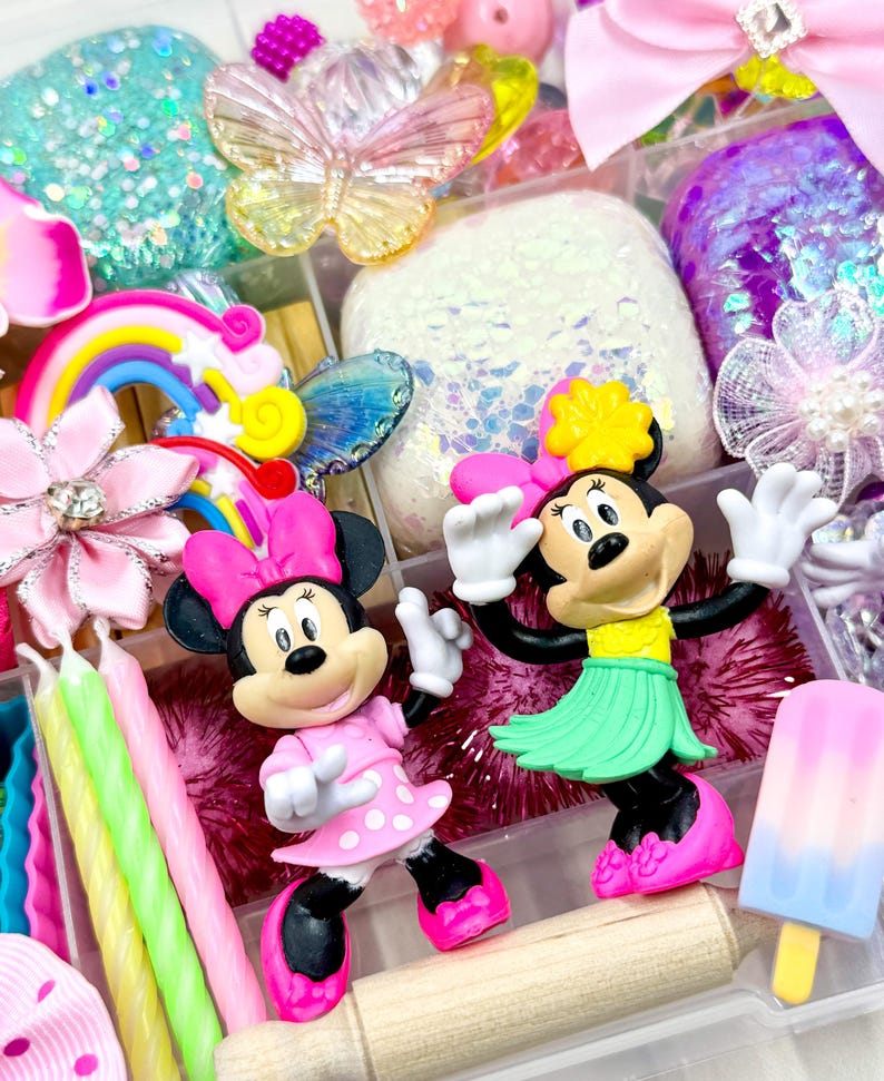 May include: A clear plastic storage box filled with colorful craft supplies, including a rainbow, a butterfly, glitter, and two small Minnie Mouse figurines. One Minnie Mouse is wearing a pink dress with white polka dots, and the other is wearing a green hula skirt.