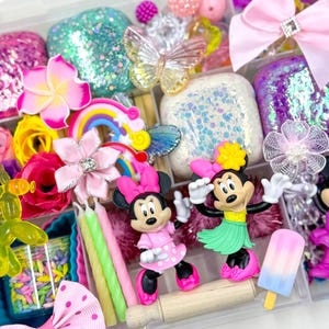 May include: A clear plastic storage box filled with various craft supplies, including colorful glitter, flowers, bows, and miniature figurines. The box also contains a rolling pin, candles, and a popsicle.