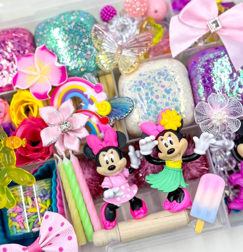 May include: A clear plastic storage box filled with various craft supplies, including colorful glitter, flowers, bows, and miniature figurines. The box also contains a rolling pin, candles, and a popsicle.