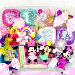 May include: A clear plastic storage box filled with colorful craft supplies, including play dough, glitter, beads, flowers, and figurines. There are three Minnie Mouse figurines, one with a hula skirt, one with a pink dress, and one with a pink dress and a bow. The box also includes a rainbow, candles, and sprinkles.