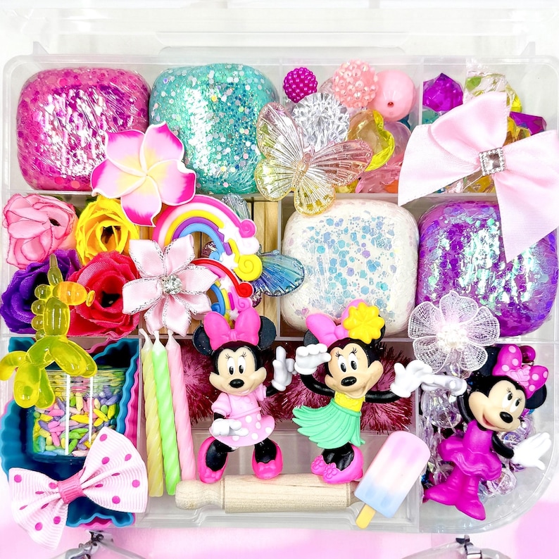 May include: A clear plastic storage box filled with colorful craft supplies, including play dough, glitter, beads, flowers, and figurines. There are three Minnie Mouse figurines, one with a hula skirt, one with a pink dress, and one with a pink dress and a bow. The box also includes a rainbow, candles, and sprinkles.