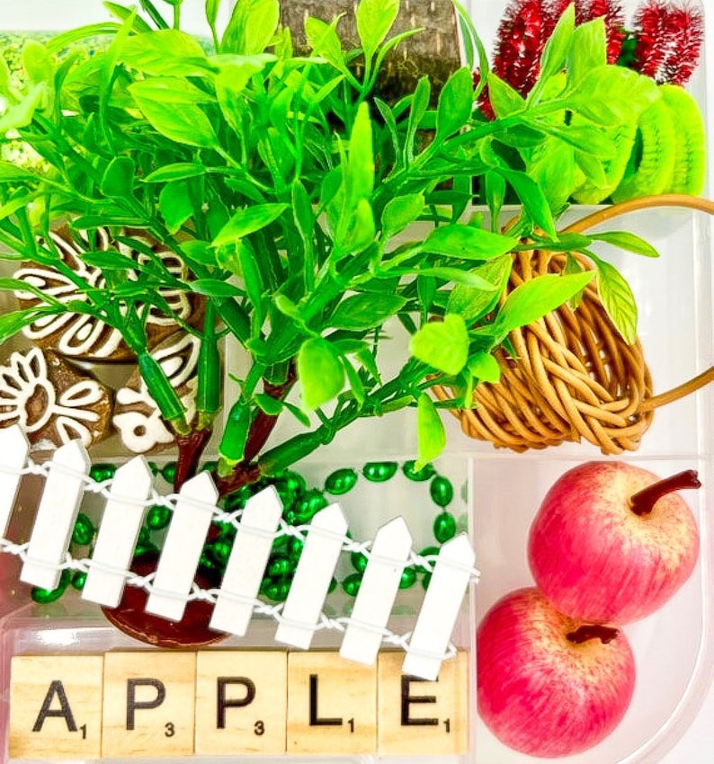 May include: A vibrant still life arrangement featuring a miniature tree with bright green leaves, a small woven basket, and two red apples. A white picket fence and the word "APPLE" spelled out with Scrabble tiles complete the scene.