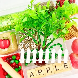 May include: A clear plastic container filled with various craft supplies. Includes a small green tree, red apples, a white picket fence, and wooden letter tiles spelling "APPLE". Other items include glitter, beads, and a rolling pin.
