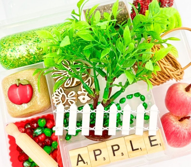May include: A clear plastic container filled with various craft supplies. Includes a small green tree, red apples, a white picket fence, and wooden letter tiles spelling "APPLE". Other items include glitter, beads, and a rolling pin.