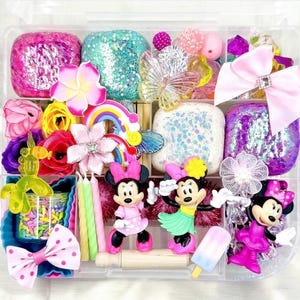 May include: A clear plastic storage box filled with colorful craft supplies, including glitter slime, rainbow sprinkles, candles, and miniature Disney characters. The box is organized into compartments with a variety of items in each.