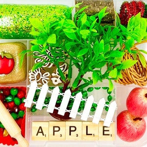 May include: A clear plastic container filled with various decorative items. Includes a green glittery rectangle, a gold square with a red apple, a miniature rolling pin, and the word "APPLE" spelled out with letter tiles. Also features a small tree and two red apples.