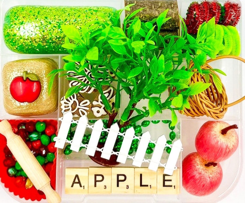 May include: A clear plastic container filled with various decorative items. Includes a green glittery rectangle, a gold square with a red apple, a miniature rolling pin, and the word "APPLE" spelled out with letter tiles. Also features a small tree and two red apples.