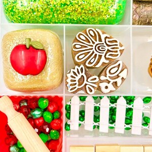 May include: A divided tray filled with various craft supplies. Includes a green glittery substance, a gold-colored square with a red apple, decorated cookies, red and green beads, a small rolling pin, and wooden letter tiles spelling "APP".