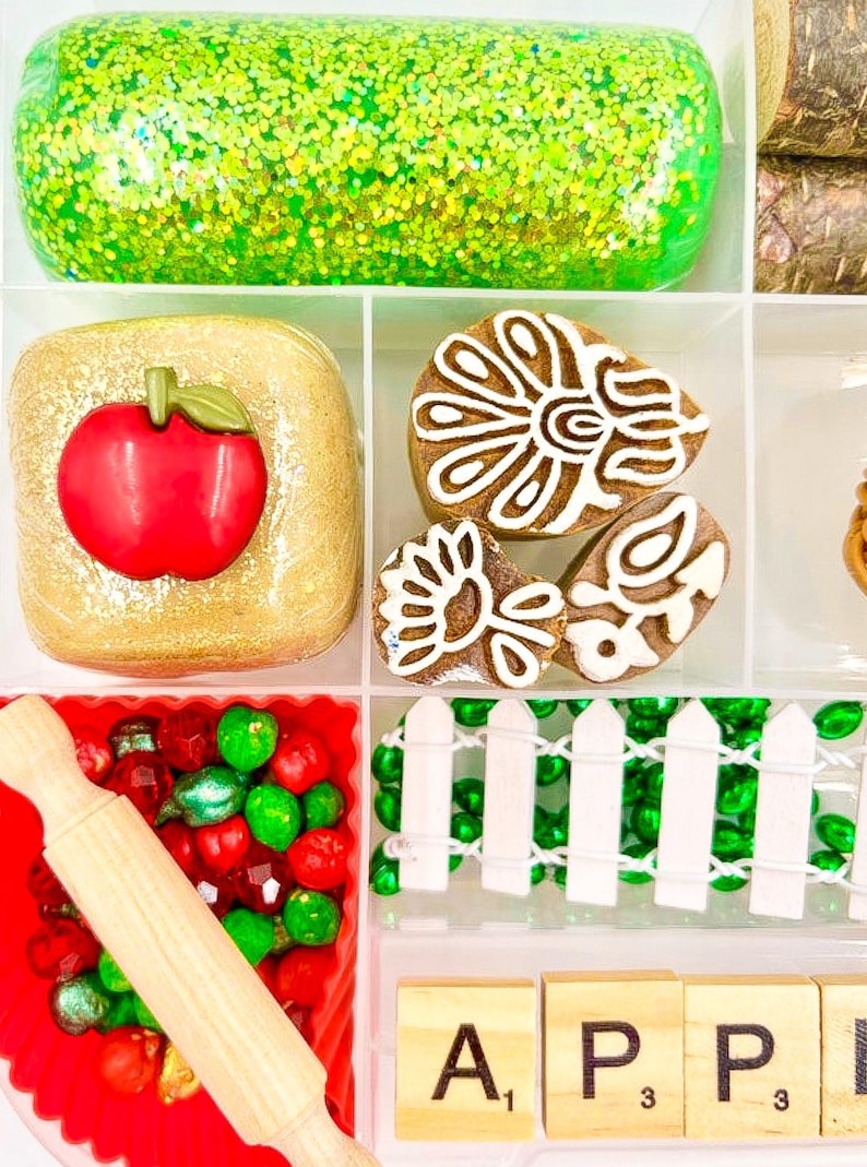 May include: A divided tray filled with various craft supplies. Includes a green glittery substance, a gold-colored square with a red apple, decorated cookies, red and green beads, a small rolling pin, and wooden letter tiles spelling "APP".