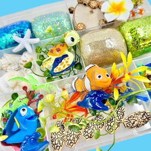 Peut inclure: Un organisateur en plastique transparent rempli de fournitures d'artisanat sur le thème de l'océan. Comprend des blocs scintillants, des étoiles de mer, des coquillages et des figurines de tortue de mer, de poisson-clown et de poisson chirurgien bleu. Éléments décoratifs : plantes artificielles et fleur blanche.