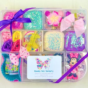 May include: A clear plastic storage box filled with various sensory items, including glitter, slime, flowers, and a small Minnie Mouse figurine. The box is tied with a purple ribbon and has a label that reads "Ready Set Sensory".