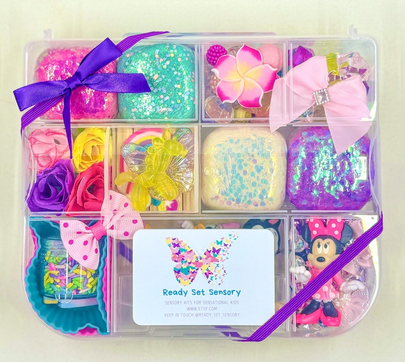 May include: A clear plastic storage box filled with various sensory items, including glitter, slime, flowers, and a small Minnie Mouse figurine. The box is tied with a purple ribbon and has a label that reads "Ready Set Sensory".