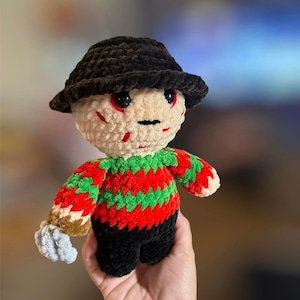 May include: Crocheted plush toy of a horror movie character wearing a black hat and a red and green striped shirt.