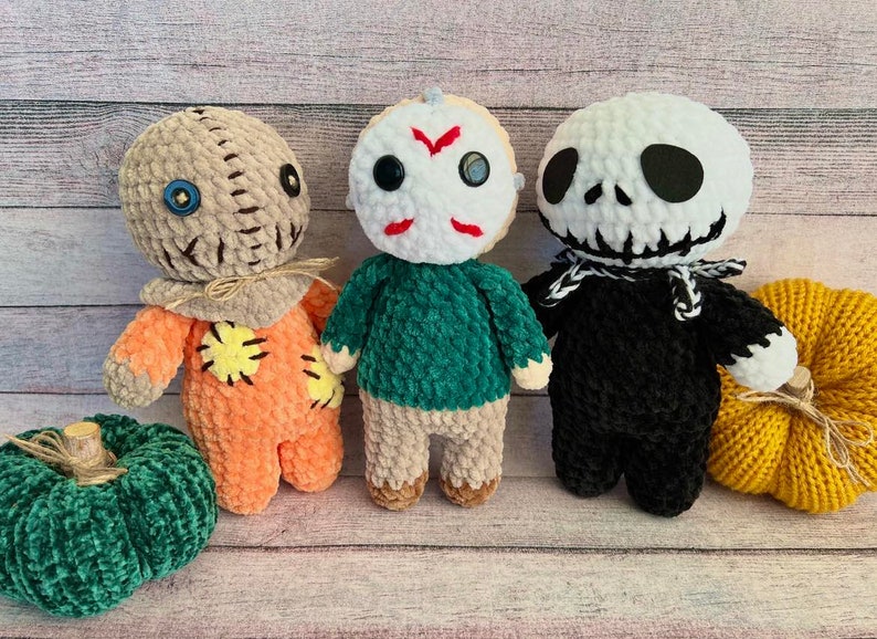 May include: Three crocheted Halloween character toys: a brown and orange scarecrow, a green and white Michael Myers, and a black and white Jack Skellington. The toys are sitting on a wooden surface with two crocheted pumpkins.