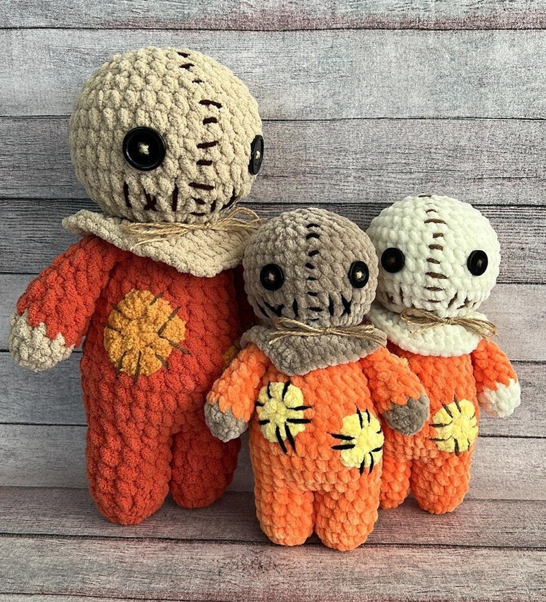 May include: Three crocheted plush toys in the shape of a character from a horror movie. The largest toy is orange with a beige head and a stitched-on mouth. The middle toy is orange with a brown head and a stitched-on mouth. The smallest toy is white with a beige head and a stitched-on mouth.