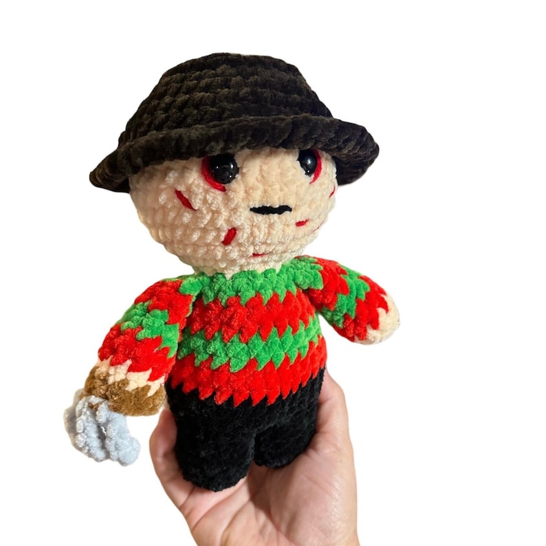 May include: A crocheted plush toy of a horror movie character wearing a black hat and a red and green striped sweater.