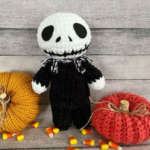 May include: A crocheted Jack Skellington doll with a white face and black body. The doll is surrounded by orange and red crocheted pumpkins and candy corn.
