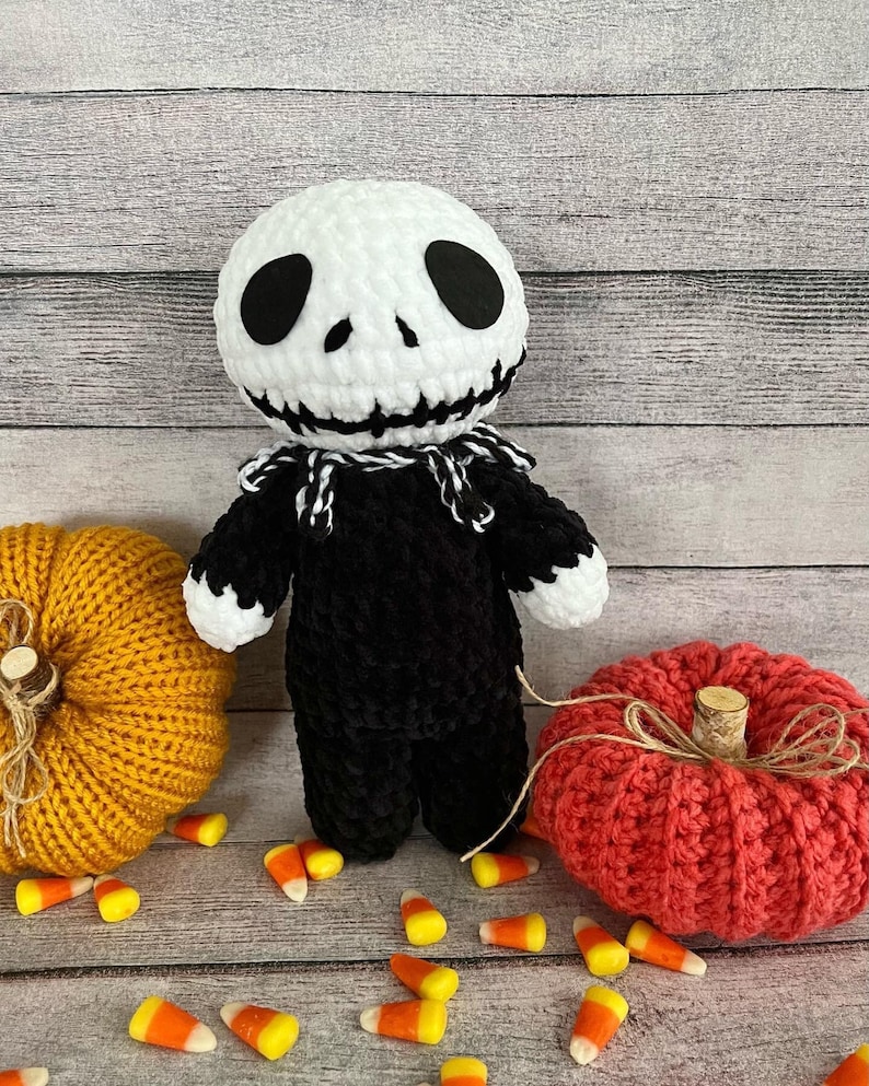 May include: A crocheted Jack Skellington doll with a white face and black body. The doll is surrounded by orange and red crocheted pumpkins and candy corn.