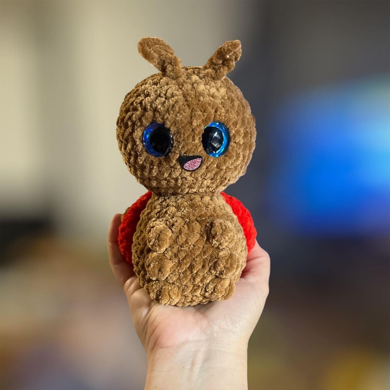 May include: A brown crocheted ladybug toy with large blue eyes and a red shell. The toy is being held in a person's hand.