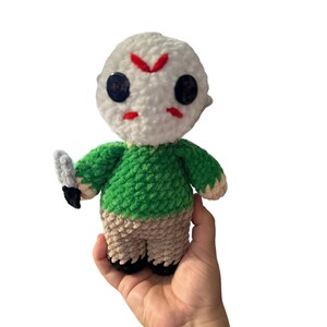May include: A crocheted plush toy of a character wearing a white mask with a red slash across the face, a green shirt, and tan pants. The toy is holding a knife in its left hand.
