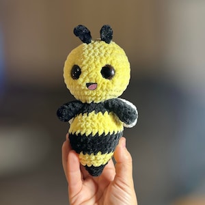 Puede incluir: Un juguete de peluche de abeja tejido a crochet con rayas negras y amarillas, ojos grandes y una boca sonriente. La abeja está en la mano de una persona.