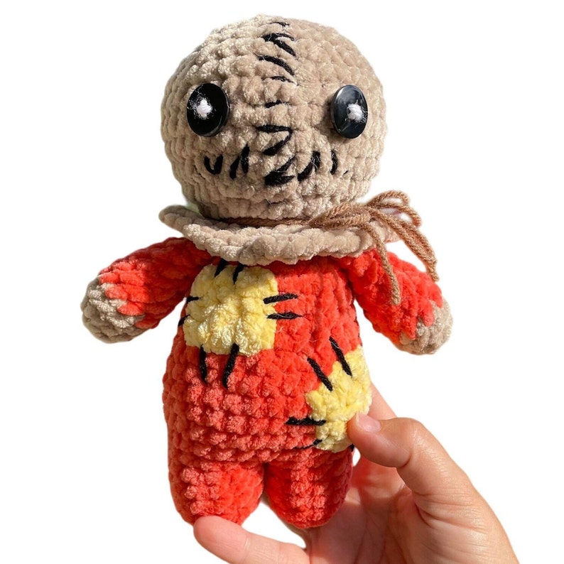 May include: A crocheted plush toy resembling a character from a horror movie. The toy is wearing a brown sackcloth mask with stitched eyes and a red and yellow patchwork outfit.