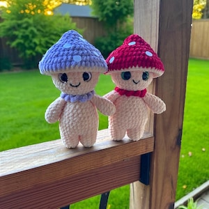 May include: Two handmade crochet mushroom figures. One has a lavender cap with white spots and a matching collar. The other has a red cap with white spots and a red collar. Both have smiling faces and small arms and legs.