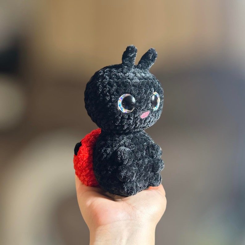 May include: A black and red crocheted ladybug plush toy with large, sparkly eyes. The ladybug has a small, pink mouth and is sitting on a hand.